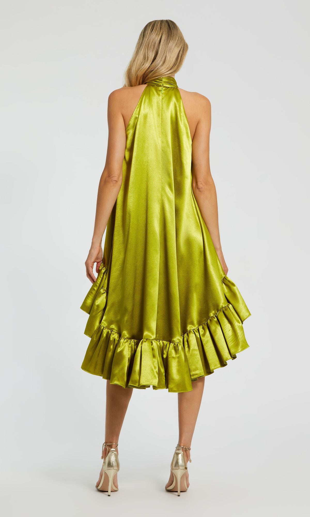 A woman with long blonde hair is seen from the back wearing the Mac Duggal 20711 satin wedding guest dress in shiny green, featuring a ruffled high-low hemline, paired with metallic high-heeled shoes.